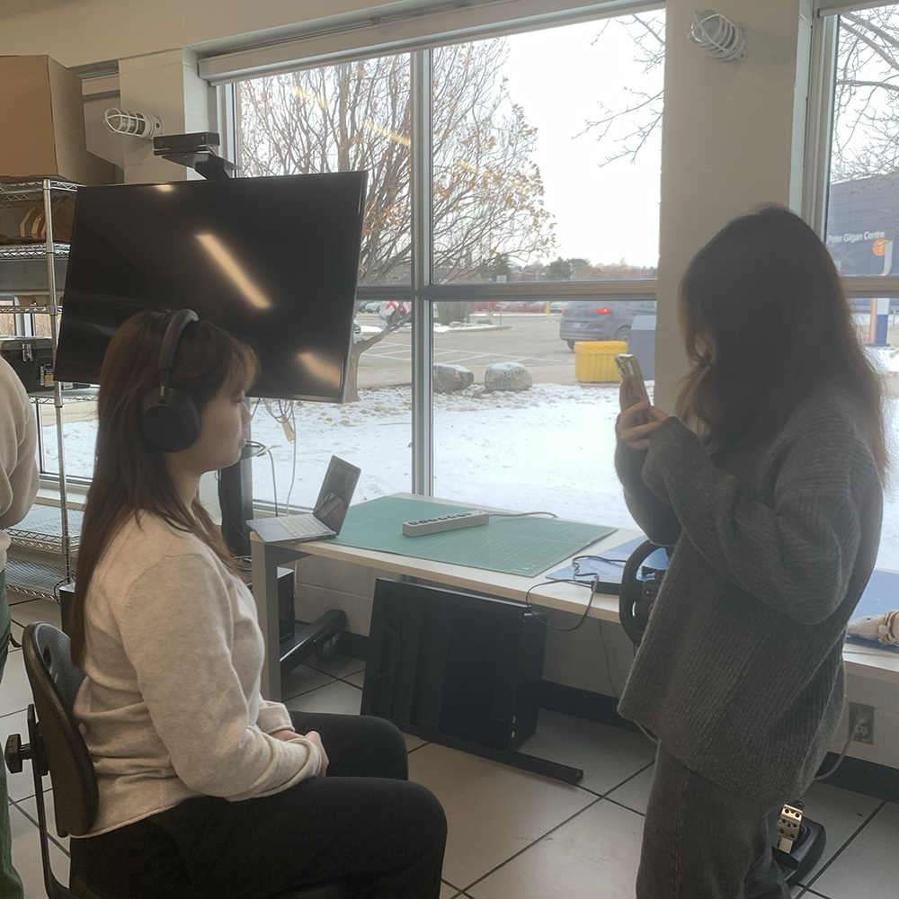 <b>Continuing the Scan</b><br> In this photo, my classmate continues to scan me, capturing different angles. I remain seated, focusing on staying still to ensure the accuracy of the 3D model we’re creating.