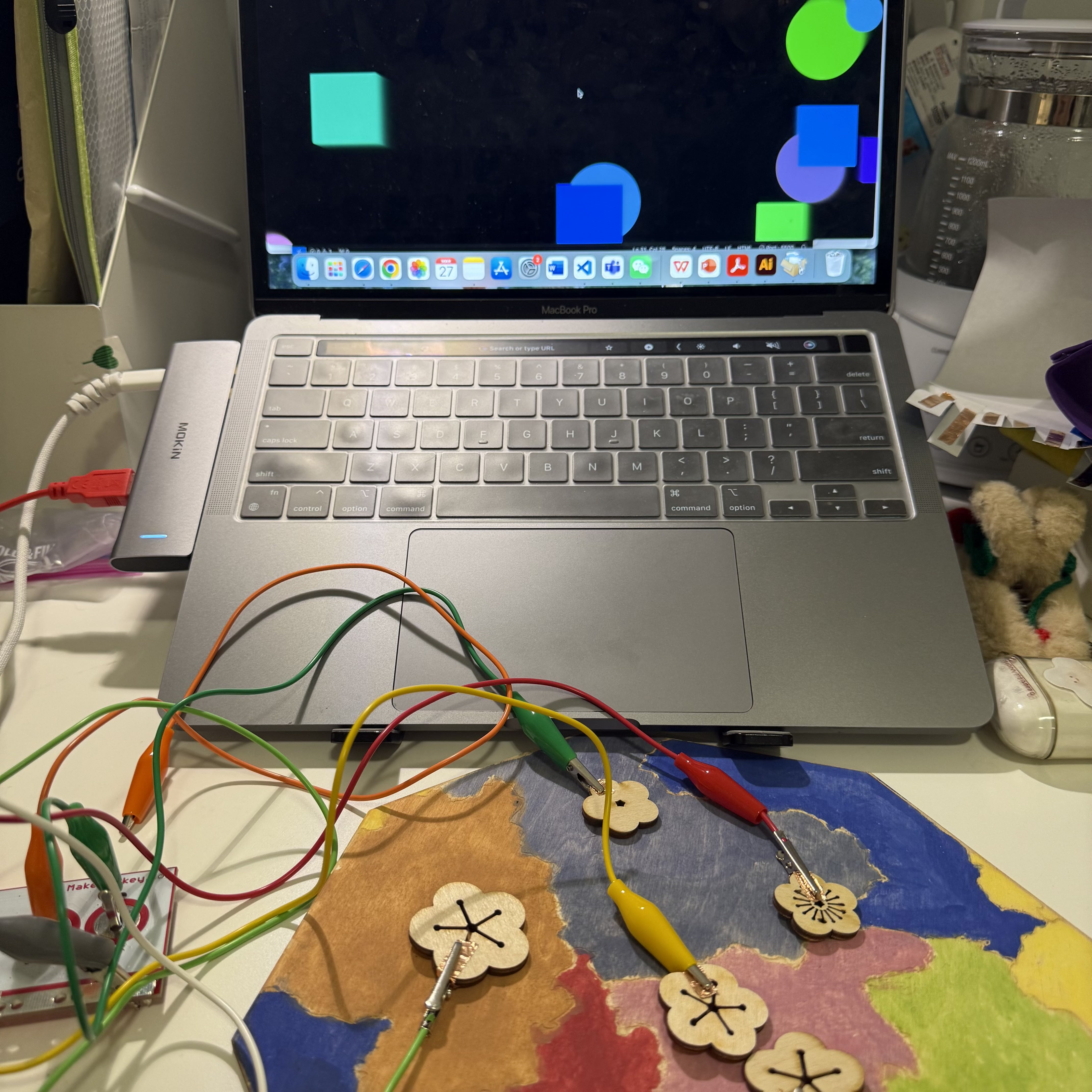 I have connected my Makey Makey board to my laptop, which displays colorful geometric shapes on the screen. I use decorative wooden flowers attached to a colorful hand-painted map as interactive buttons. Each flower is connected to the Makey Makey with alligator clips, turning them into touch-sensitive controls.