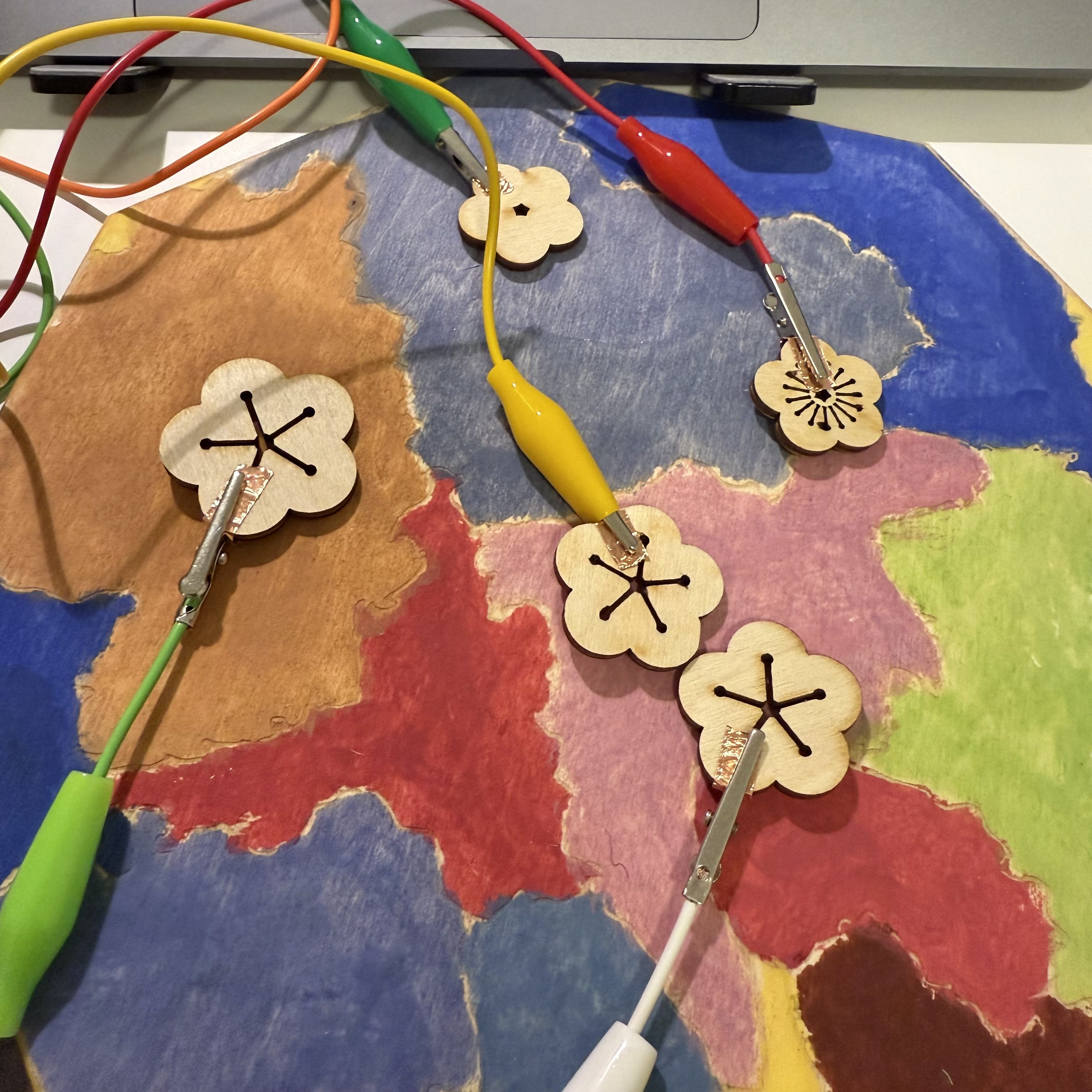 In this close-up, you can see the wooden flowers placed on a hand-painted map. Each flower is connected to the Makey Makey via alligator clips, which are attached to small pieces of copper tape on the back of each flower. This setup allows me to interact with digital programs by simply touching these flowers.
